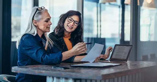 Two women discussing documents at a table with a laptop.