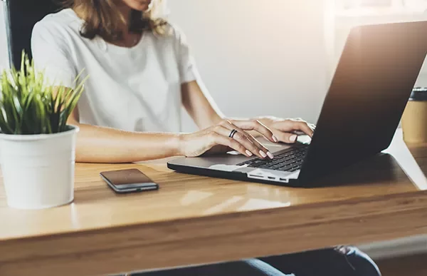 Person typing on a laptop at a wooden desk with a plant and smartphone nearby.