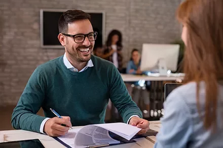 Smiling man in a green sweater holds papers while talking to a woman in an office setting.